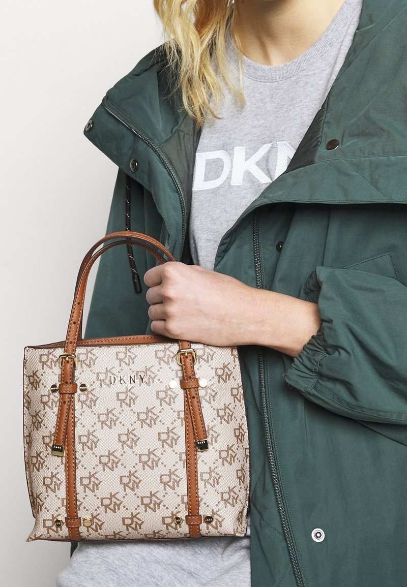 Kvinde iført grøn jakke og grå DKNY skjorte, der holder en beige og brun DKNY monogram taske ved håndtagene.