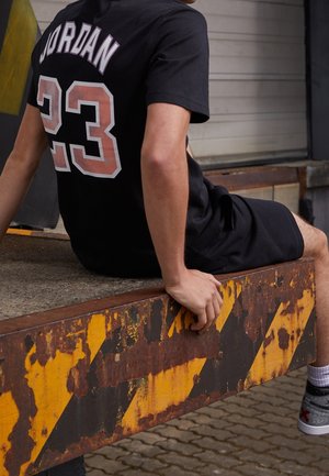 Sort t-shirt med hvid tekst "JORDAN" og "23" på ryggen; siddende på en rusten, gulstribet metaloverflade, iført sorte shorts og sneakers.
