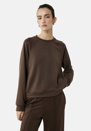 Vrouw met donker haar die een bruine trui met lange mouwen en bijpassende broek draagt, staand met één hand in de zak op een lichte achtergrond.