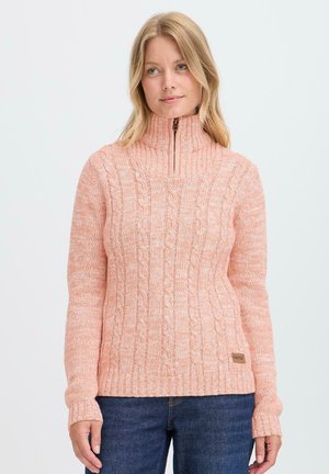 Vrouw in een perzik kleurige kabeltrui met een halve ritssluiting en hoge kraag, draagt een spijkerbroek tegen een lichtgekleurde achtergrond.