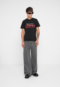 Black t-shirt with red text reading "1981 REPLAY," paired with loose gray jeans and black platform shoes. Accessories include black sunglasses.