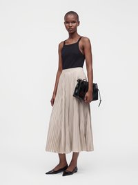 Femme portant un haut noir sans manches, une jupe midi plissée beige, des ballerines noires pointues, tenant un sac à main noir, debout devant un fond uni.