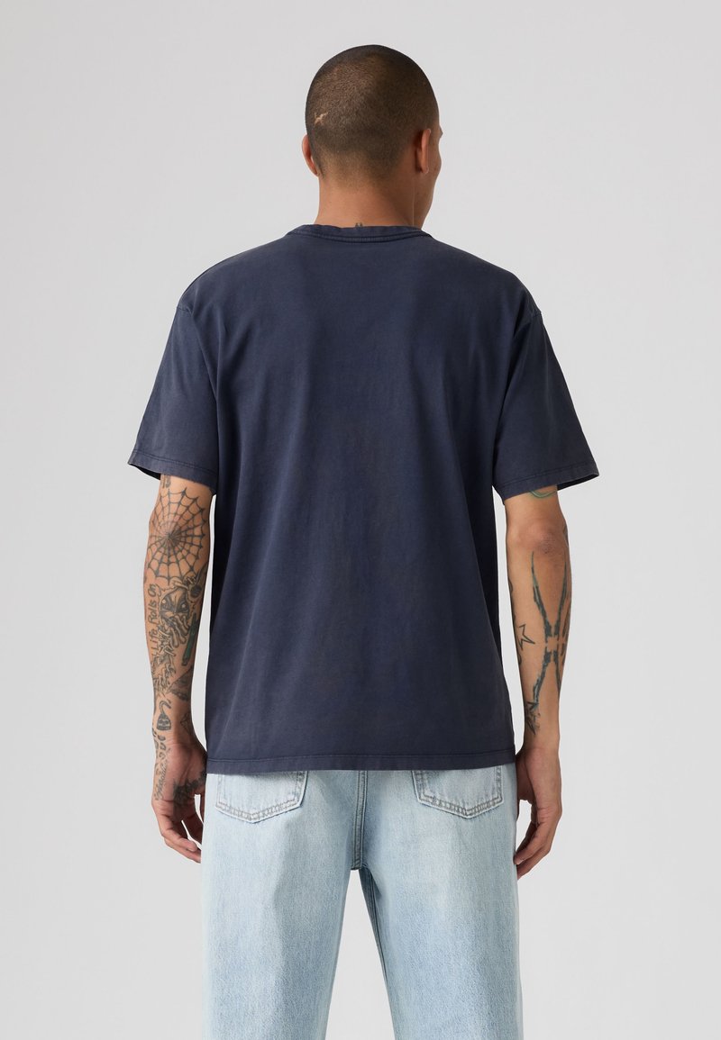 Uomo con le braccia tatuate indossa una maglietta blu scuro a maniche corte e jeans azzurri, in piedi di spalle su uno sfondo semplice.