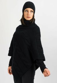 Svart stickad poncho med kabelmönster, hög krage och ribbad trim, i kombination med en matchande mössa, vilket ger en texturerad och lager-på-lager-look.