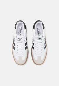 Pár bílých tenisek Adidas s černými pruhy, béžovými semišovými špičkami, gumovými podrážkami a bílými tkaničkami, zobrazených shora na bílém pozadí.