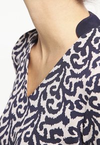 Nærbillede af en persons nakke og øvre bryst iført et sort og hvidt abstrakt mønstret V-udskåret tøj med en mørk kraveflige.