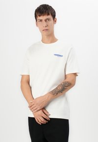 Ung mann med kort mørkt hår iført en hvit "UNDERDOGS"-t-skjorte og svarte bukser, som viser et tatovering av roser og en klokke på venstre underarm.