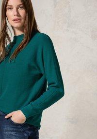 Teal ribbad stickad tröja med hög hals och långa ärmar, med en avslappnad passform. Buren över mörk denim med en lätt blekning.