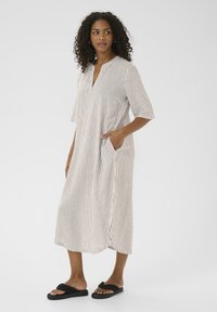 Abito midi a righe in crema e marrone, con scollatura a notch, maniche corte, tasche laterali e vestibilità rilassata. Indossato con sandali neri.