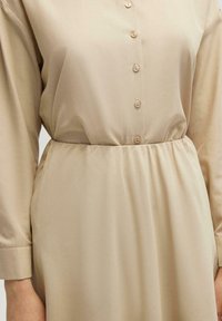 Robe beige avec un devant boutonné, des manches longues avec un poignet, et une taille élastique pour un ajustement décontracté. Tissu lisse avec un léger brillant.