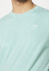 Světle mátová tričko vyrobené z měkké bavlny, s hladkou texturou a jemným vybledlým vzorem. Obsahuje malé bílé logo Nike na hrudi.