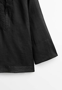 Top en lin noir à coupe ample, avec un col en V, une fermeture à lacet et des manches longues. Le tissu a une texture lisse et légèrement froissée.