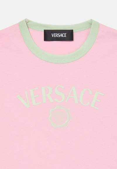 Rozā kokvilnas t-krekls ar gaiši zaļu ribotu apkakli, kurā ir izšūts "VERSACE" teksts un logotipa detaļa centrā.