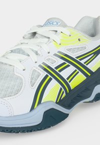 Λευκό και γκρι αθλητικό παπούτσι Asics με κίτρινα και μαύρα ριγέ στοιχεία και λευκά κορδόνια σε στιβαρή σκούρα σόλα.