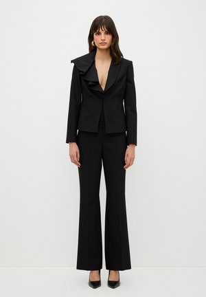Completo nero su misura con colletto arricciato, giacca slim-fit e pantaloni a zampa. Tessuto liscio con finitura opaca. Scarpe con tacco alto lucide.