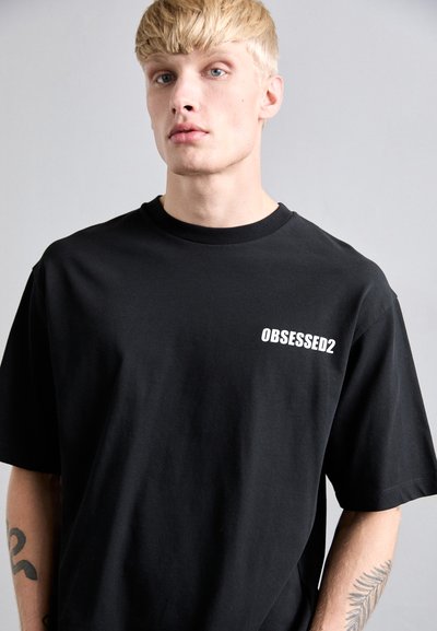 Uomo biondo con pelle chiara che indossa una maglietta nera con il testo bianco "OBSESSED2", mostrando tatuaggi su entrambi i avambracci su uno sfondo semplice.