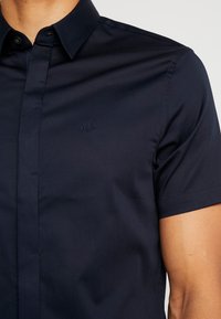 Modrá košile s krátkým rukávem vyrobená z hladké tkaniny s límečkem na knoflíky. Obsahuje tonální logo na hrudi a má slim fit střih.