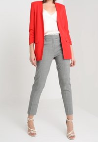 Blazer rouge, blouse blanche, pantalon à carreaux gris, et talons blancs à lanières. Le blazer a des manches longues, une coupe ajustée et un design ouvert à l'avant.