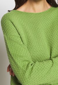 Groen gebreide trui met een oversized halslijn en lange mouwen. De stof heeft een diamantpatroon, wat diepte en uitstraling toevoegt.