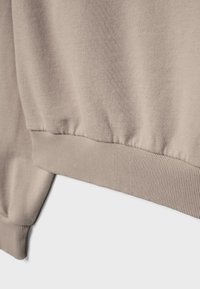 Beige sweatshirtstof med rynket ribkant for neden og ved ærmet, der viser tekstur og syningsdetaljer på en ensfarvet baggrund.