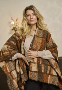 Poncho à carreaux marron et crème en matériau texturé, avec un bord à franges, porté sur un haut beige avec des manches bouffantes.