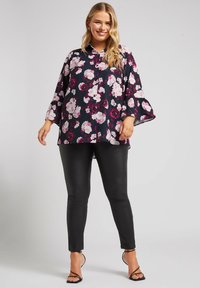 Blusa nera floreale con fiori rosa e bianchi, maniche a campana e colletto, abbinata a pantaloni neri aderenti e tacchi neri con cinturini.