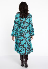 Langärmliges Kleid mit türkisem Blumenmuster auf schwarzem Hintergrund, mit Rüschensleeves und einer fließenden Silhouette. Getragen mit kniehohen Stiefeln.