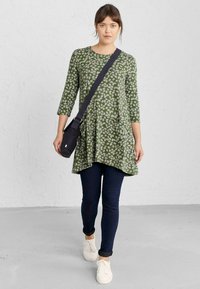 Zelena tunika s bijelim uzorkom lišća, tričetvrt rukavi, uparen sa tamnim uskim trapericama i bijelim tenisicama, nosi malu crnu torbicu.