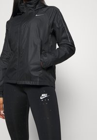 Czarna kurtka Nike z wysokim kołnierzem, zapięciem na zamek i odblaskowymi akcentami. W zestawie z czarnymi legginsami z białym logo Nike.