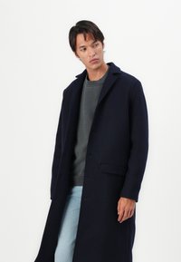 Cappotto lungo navy con design monopetto, caratterizzato da un colletto a cappotto, tasche frontali e una finitura pulita. Indossato sopra un maglione grigio.