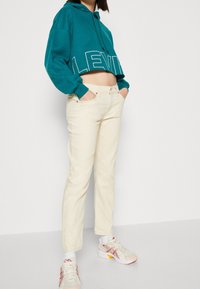 Sweat à capuche court bleu sarcelle avec lettrage blanc « LEVI'S », associé à un jean droit couleur crème et des baskets athlétiques aux tons neutres.