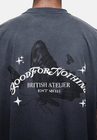 Czarny t-shirt z ciemnoszarym odcieniem, z dużym motywem motyla na plecach, z białym napisem: "NIE PRZYDATNE BRYTYJSKIE ATELIER ZAŁ. 2011."