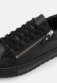 Schwarze Leder-Sneaker mit flachen schwarzen Schnürsenkeln, einem seitlichen Reißverschluss mit metallischen Details und einer strukturierten Gummisohle.