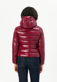 Bordeaux pufferjack met een glanzende afwerking, horizontale quilt-stiksels en een ronde capuchon. Gecombineerd met donkere denim jeans, gezien vanuit de achterkant.