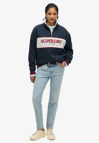 Sudadera de cuello con cremallera en navy con una franja blanca y el logo rojo "SUPERDRY", combinada con jeans rectos de pierna ligera en azul claro y zapatillas crema.
