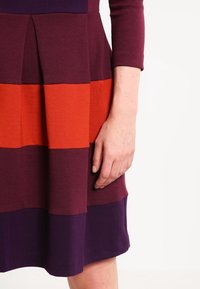 Sukienka z poziomymi paskami w głębokim fioletowym, bordowym i pomarańczowym kolorze. Materiał ma teksturowane wykończenie. Wzór obejmuje plisowany dół.