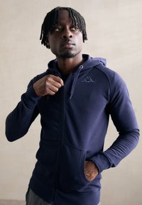 Námornícky modrá mikina na zips vyrobená z mäkkej látky, s prednou vreckom, kapucňou so sťahovacou šnúrkou a malým logom na hrudi.
