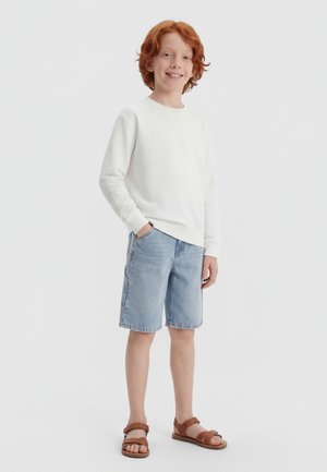 Rudy chłopiec uśmiechający się, ubrany w białą koszulę z długim rękawem, jasnoniebieskie krótkie spodenki dżinsowe i brązowe sandały, stojący z jedną ręką w kieszeni.