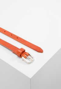 Oranž nahast vöö tekstureeritud viimistlusega, millel on hõbedane metallpannal ja mitu aukude rida. Laotatud tasasele valgele pinnale.