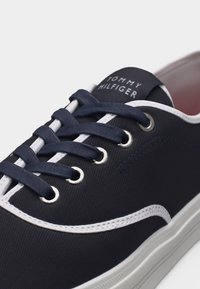 Gros plan sur une chaussure de sport bleu marine avec des surpiqûres blanches, des œillets métalliques, des lacets bleu marine et une étiquette sur la languette indiquant "Tommy Hilfiger".