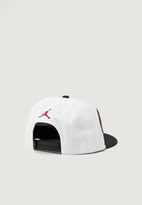 Bijela kapa sa ravnim vizir ocrnjena, s crvenim izvezenim Jumpman logotipom na poleđini i podesivim plastičnim kopčanjem.