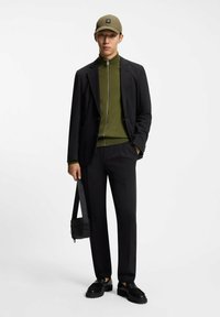 Abito nero su maglione verde con cerniera, abbinato a un berretto verde e scarpe nere. Una piccola borsa nera è appesa a una spalla.