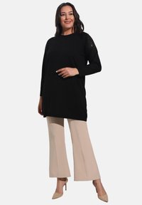 Black long-sleeve tunic with textured shoulders, paired with beige flared pants and nude heels, against a plain white background.