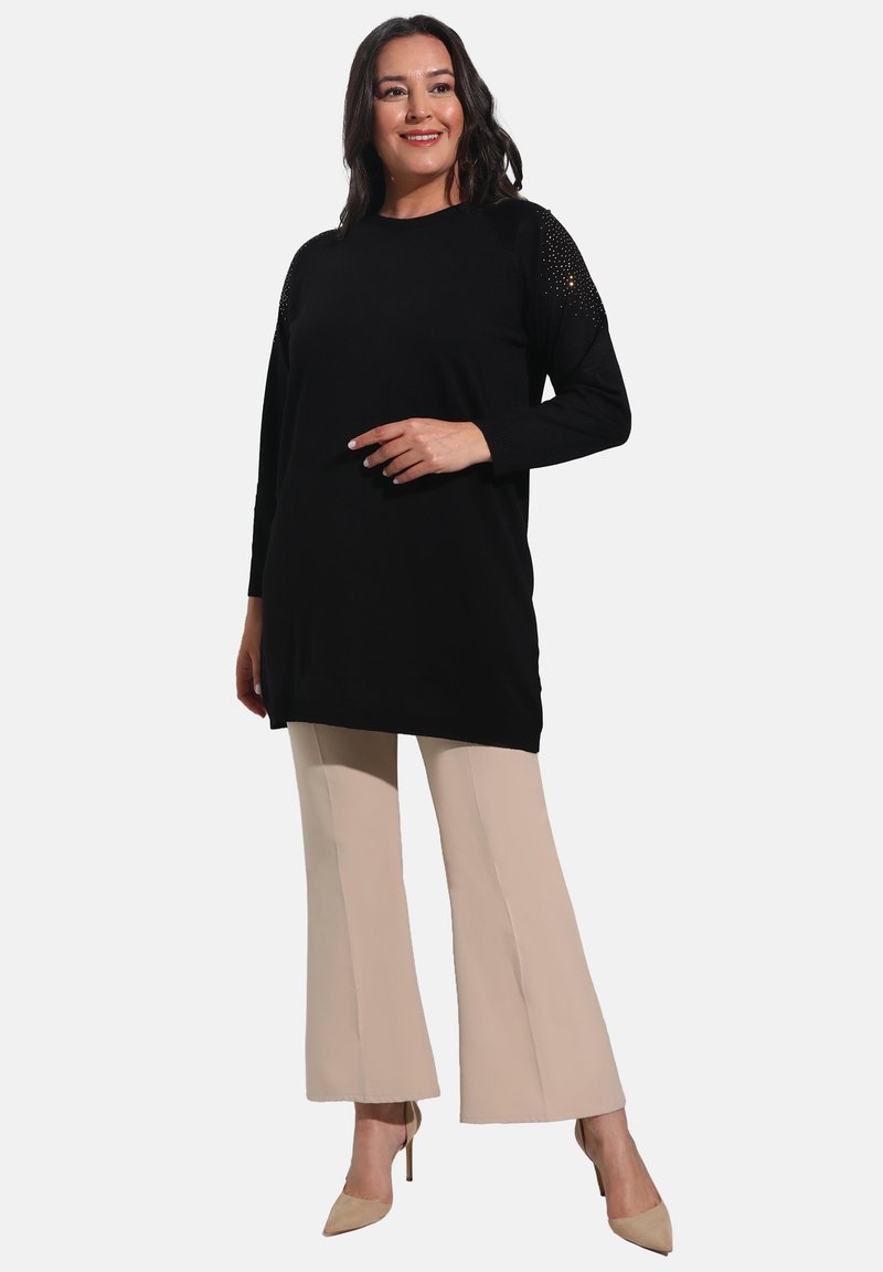 Black long-sleeve tunic with textured shoulders, paired with beige flared pants and nude heels, against a plain white background.