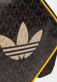 Hnědá kožená taška s vzorovaným pozadím překrývajících se listových tvarů. Výrazné logo ve smetanové barvě s pruhy dodává kontrast, okraje jsou žluté.