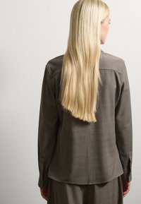 Donna con lunghi capelli biondi lisci, che indossa una maglia marrone larga e pantaloni coordinati, in piedi di fronte a un muro semplice di colore chiaro.