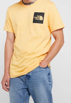 Mężczyzna w żółtej koszulce The North Face i niebieskich dżinsach, z jedną ręką w kieszeni, pokazujący tatuaż na szyi i części ramienia.