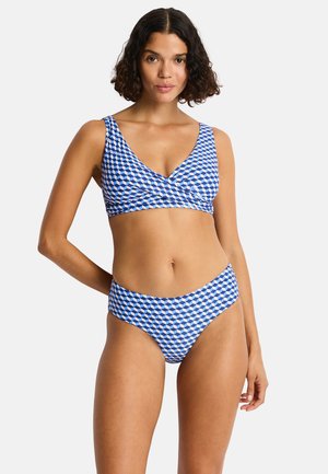 Donna che indossa un bikini con motivo geometrico blu, bianco e nero, con top a scollo a V e slip a vita alta, in piedi su uno sfondo semplice.