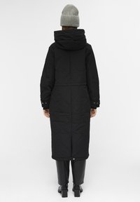 Person iført en lang sort quiltet jakke med hætte, sorte bukser, sorte hæle støvler og en grå strikbeanie, stående med ryggen til.