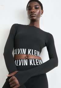 Selbstbewusste Frau in einem schwarzen Langarm-Crop-Top und passenden Leggings mit weißen "Calvin Klein" Logo-Bändern, die vor einem weißen Hintergrund posiert.